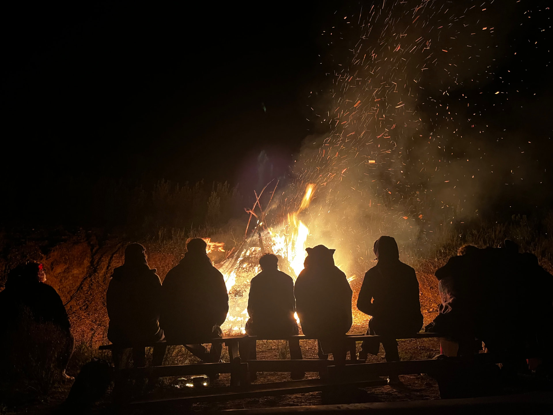 People sitting in silhouette on a bench in front of a bonfire at night